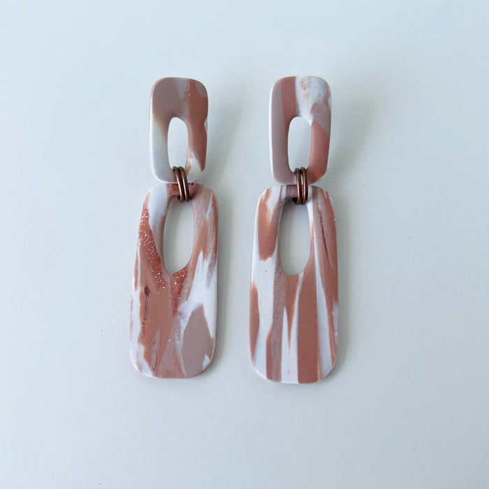A pair of handmade rectangular drop earrings with a cutout center, made from marbled polymer clay in shades of terracotta brown, shimmering copper, and white. The top and bottom segments are joined with antique copper-colored jump rings. The earrings have embedded titanium posts.