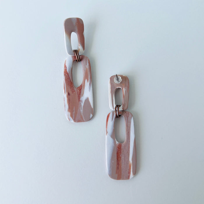 A pair of handmade rectangular drop earrings with a cutout center, made from marbled polymer clay in shades of terracotta brown, shimmering copper, and white. The top and bottom segments are joined with antique copper-colored jump rings. The earrings have embedded titanium posts.