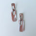 A pair of handmade rectangular drop earrings with a cutout center, made from marbled polymer clay in shades of terracotta brown, shimmering copper, and white. The top and bottom segments are joined with antique copper-colored jump rings. The earrings have embedded titanium posts.