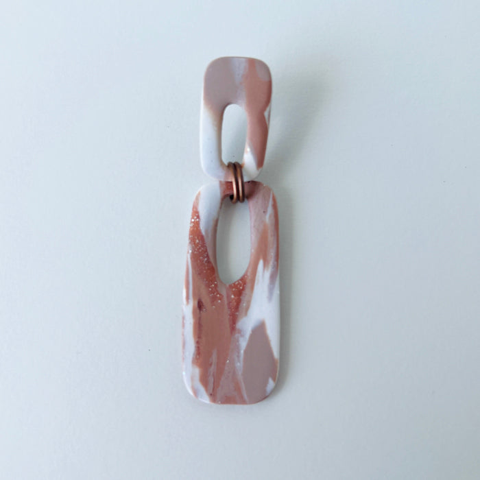 A handmade rectangular drop earring with a cutout center, made from marbled polymer clay in shades of terracotta brown, shimmering copper, and white. The top and bottom segments are joined with antique copper-colored jump rings. The earrings have embedded titanium posts.