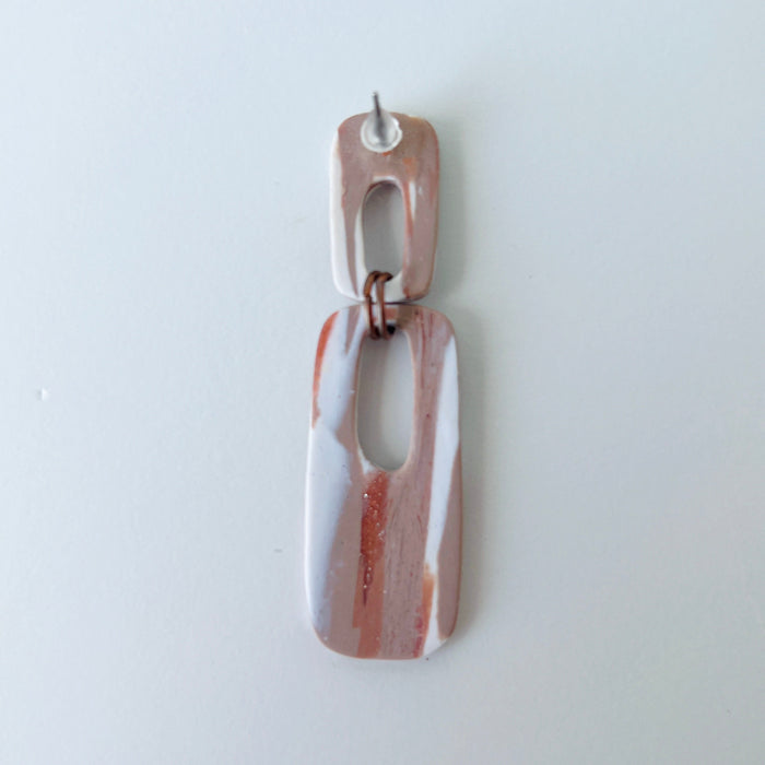A handmade rectangular drop earring with a cutout center, made from marbled polymer clay in shades of terracotta brown, shimmering copper, and white. The top and bottom segments are joined with antique copper-colored jump rings. The earrings have embedded titanium posts.