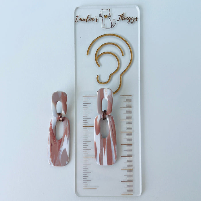 A pair of handmade rectangular drop earrings with a cutout center, made from marbled polymer clay in shades of terracotta brown, shimmering copper, and white. The top and bottom segments are joined with antique copper-colored jump rings. The earrings have embedded titanium posts.