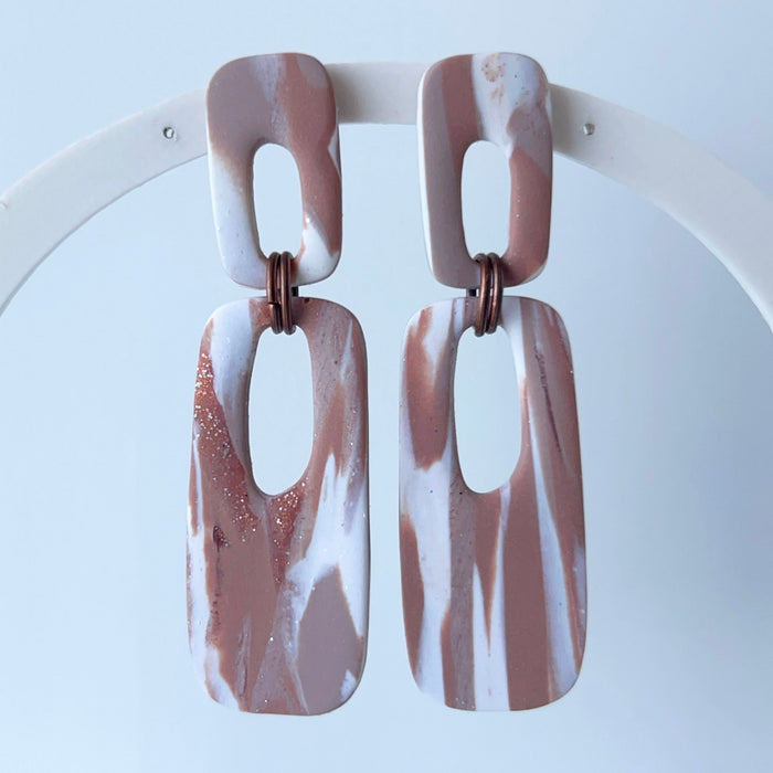 A pair of handmade rectangular drop earrings on a hanging white display arch.  The rectangular drop earrings have a cutout center, made from marbled polymer clay in shades of terracotta brown, shimmering copper, and white. The top and bottom segments are joined with antique copper-colored jump rings. The earrings have embedded titanium posts.