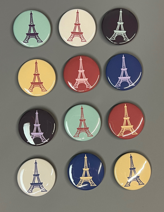 A gray background has twelve round shiny magnets with line drawings of the Eiffel Tower. They are in muted colors.