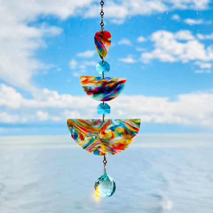 Golden Swirl: Swirled warm-toned polymer clay teardrop and arches with aqua glass crystals and a gold chain, photographed against a bright sky over water.