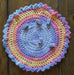 Photograph of a small, round, crocheted altar cloth with ruffled edging. It is made with pastel blue, pink, yellow, and orange multicolored yarn. There is a set of blue knucklebone dice strewn across it.