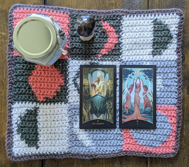 Photograph of a tarot cloth with a checkerboard moon phase pattern made with white, pink, grey, and olive green yarns and a brown border. There are two tarot cards, a candle, and a ceramic mushroom on it.
