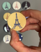 A hand holds a round magnet in light yellow with a dark blue graphic of the Eiffel Tower. The magnet is 1.5 inches across.