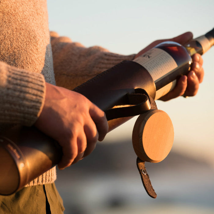 A man in a tweed sweater is pulling a bottle of Oban scotch whisky out of a handcrafted, hand-stitched leather tube case with an attached wood lid at sunset on the beach