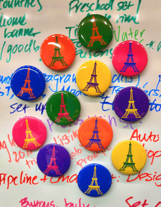 A white board has 12 round, shiny magnets in bright colors. Each has the same line drawing of the Eiffel Tower.