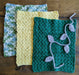 Photograph of a set of three green, yellow, and light blue crocheted kitchen towels overlapping each other, with their grey, leafy crochet tie laid across them.
