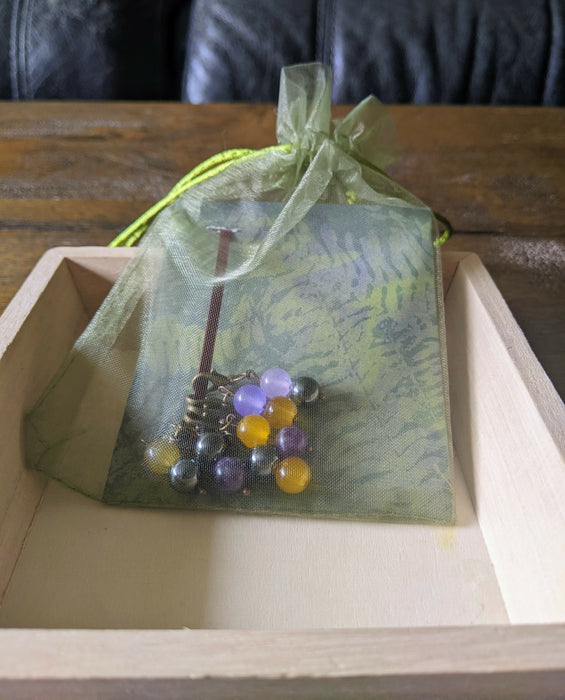 Photograph of the nonbinary stitch markers in a green tulle bag, resting in a square wooden tray.
