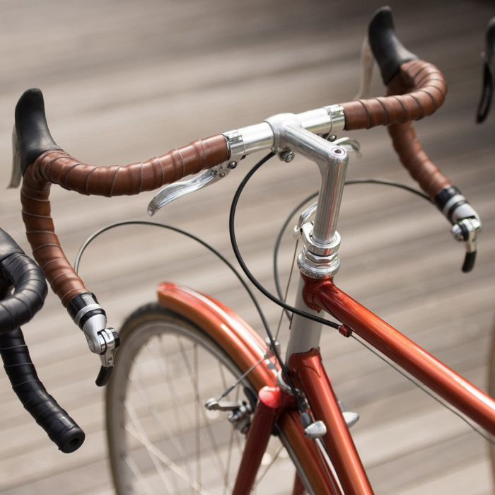 A bird's eye view looking down on three bicycles lined up in a row over a boardwalk floor. Each bicycle has a different color leather wrapped in coils on its handlebars: Dark Brown, Black and Honey color.