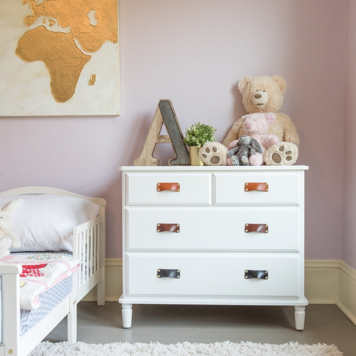 Children's nursery with natural leather bin pulls on clean white furniture. A large teddy bear sits on top of the chest of drawers and the walls are painted a pastel purple with gold accents. 