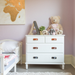 Children's nursery with natural leather bin pulls on clean white furniture. A large teddy bear sits on top of the chest of drawers and the walls are painted a pastel purple with gold accents. 