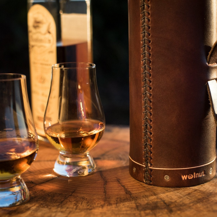 Close-up detail of the leather whiskey bottle case showing the bottom half of the case. Around the bottom of the case is a grooved out line showing the natural color of the leather below the dark brown hand-dyed surface and the "Walnut" logo.